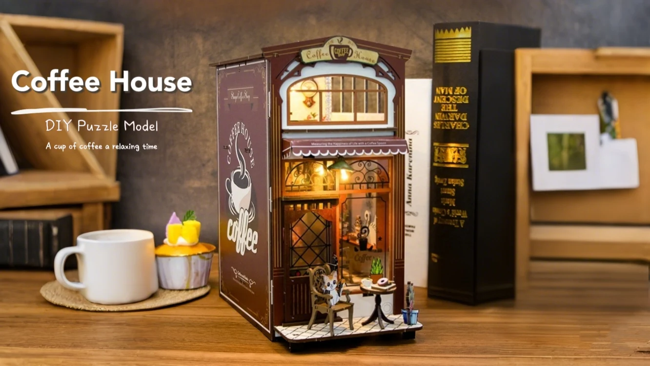 A miniature coffee house DIY puzzle model is displayed on a wooden surface. The model is accompanied by a cup of coffee and a cupcake. In the background, there are books and a wooden shelf.