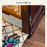 A close - up view of the coffee house DIY book nook kit, highlighting the touch - sensitive light - up feature. The image shows a part of the kit's exterior with a light - up indicator and a decorative element on the floor.