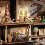 A close - up of a DIY miniature shelf in a magic wand shop, featuring a golden magic wand, a lit lantern, scrolls, and envelopes.