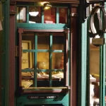 A close - up of the entrance to a DIY magic wand shop miniature. Features a green door with a window, brass - colored hardware, and "Magic Wand" signs above and below the window.