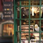 A close - up of a DIY magic wand shop miniature's window. Features "Magic Wand" signage, floating books, an owl figurine, and wands inside.