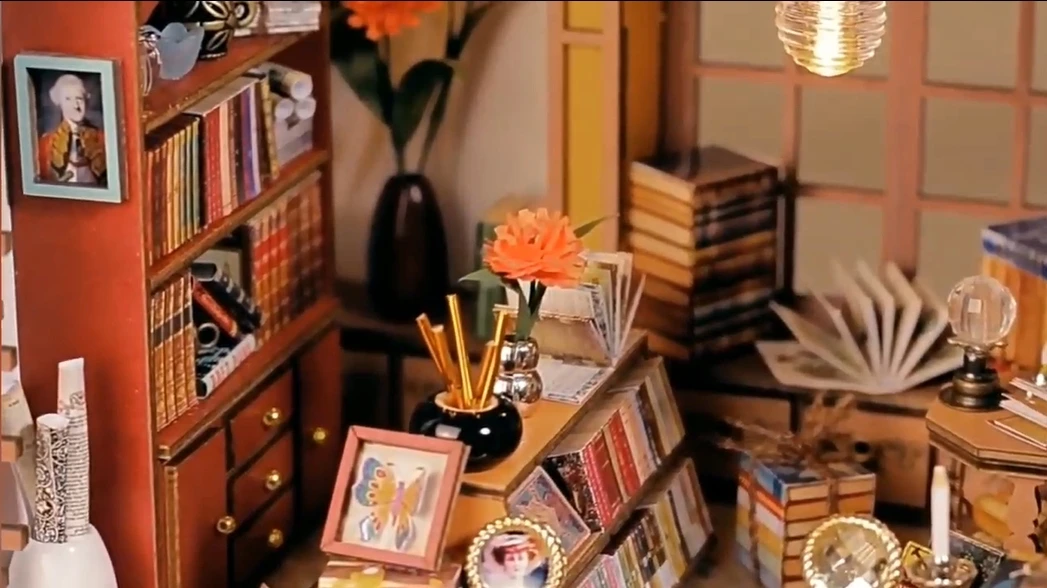A close - up of a section in a DIY magic book store miniature house, featuring bookshelves, floral decorations, framed pictures, and writing utensils.