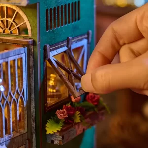 A hand opening a miniature window on the side of the Magic Shop Book Nook Kit, revealing interior details and small decorative flowers.