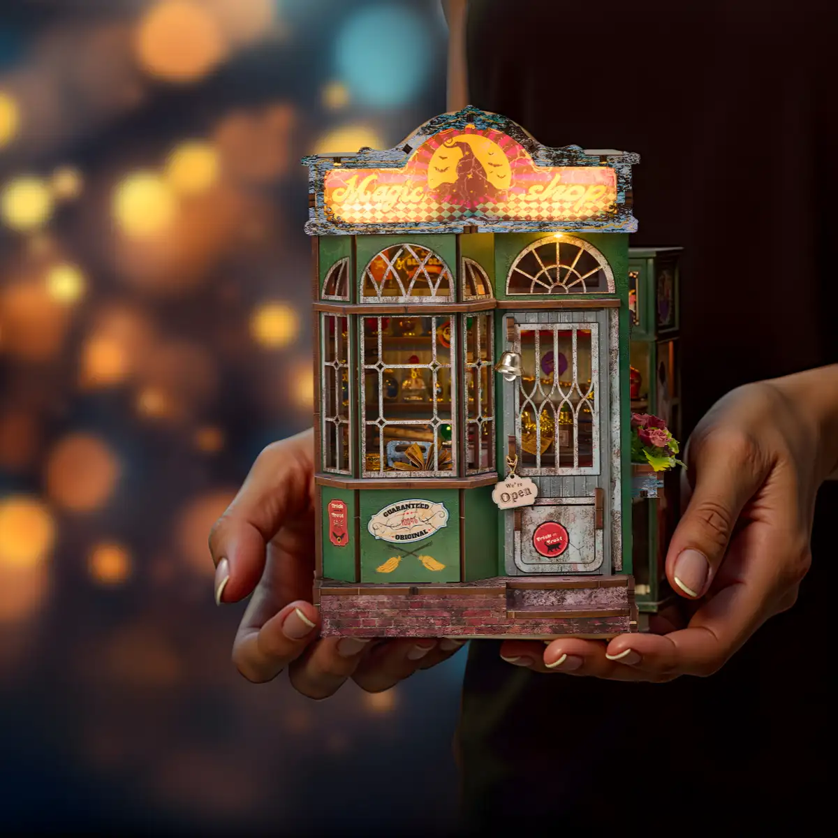 Two hands holding the fully assembled and illuminated Magic Shop Book Nook Kit against a blurred background of warm, bokeh lights.