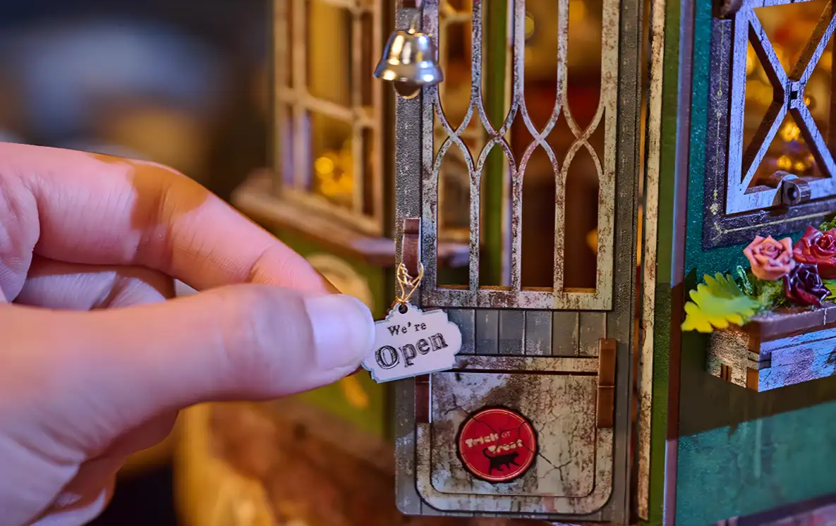 A hand hanging a tiny "We're Open" sign on the front door of the Magic Shop Book Nook Kit, which also has a small bell.