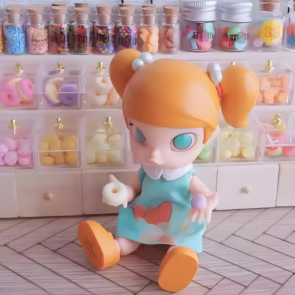 A doll holding a miniature donut and macaron, sitting on a wooden floor in front of a shelf filled with miniature sweets.