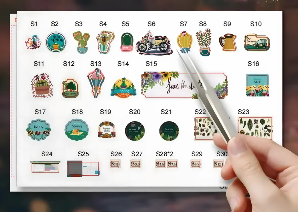 An image displaying various paper sticker accessories for the Bloomsomary Atelier flower shop book nook model, with a hand using tweezers to pick up one of the stickers.