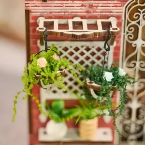 Two miniature hanging flower baskets with green foliage and small pink and white flowers, suspended from a decorative wooden structure on a red brick miniature building, with a lattice window behind.