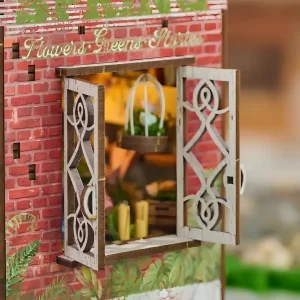 A detailed close-up of an open white wooden window on a red brick miniature building, with "Flowers Greens Stories" text above, and a tiny hanging birdcage visible inside.