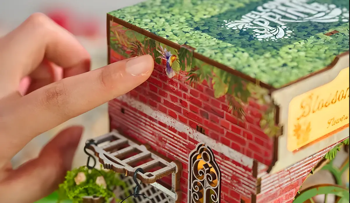 A close-up of a hand with a finger pointing at or touching a small, colorful bird sticker on the red brick wall of the Bloomsomary Atelier miniature shop, highlighting the touch-activated lighted book nook kit for collectors.