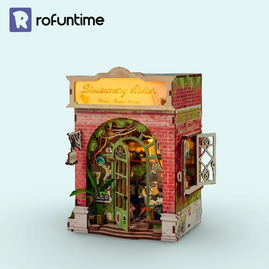 A miniature Bloomsomary Atelier flower shop book nook, lit from within, standing on a light blue background. The shop features red brick walls, a green arched entrance, and a glowing sign.