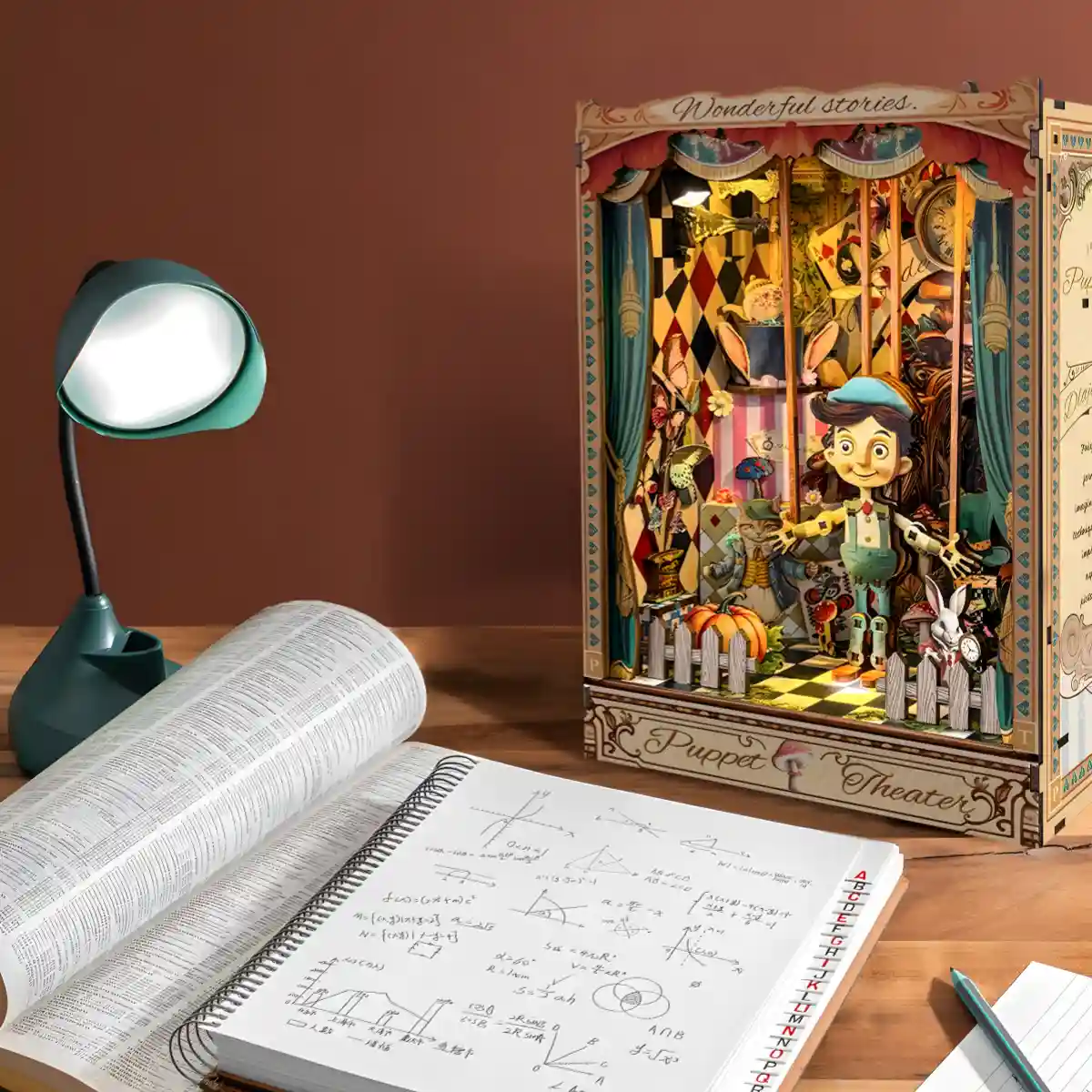 The lit Puppet Theater Book Nook Kit on a wooden desk with an open textbook and a green desk lamp.