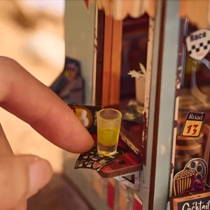 A finger pointing to a tiny yellow drink in a glass on a miniature outdoor counter of the book nook kit.