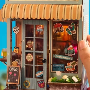 A hand touching the side of a miniature beach bar book nook kit, showing detailed windows, door, and exterior decorations.