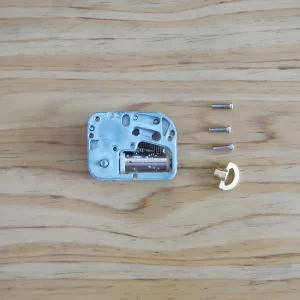 A close-up view of the internal mechanism of a mini music box, with three small screws and a gold-colored winding key laid out beside it on a wooden surface.