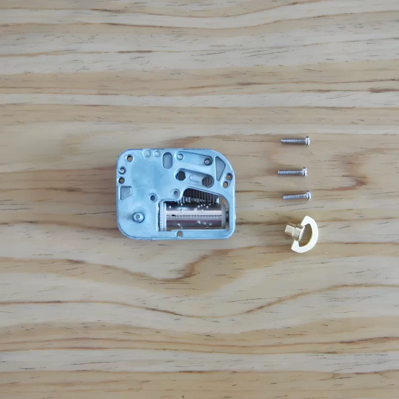 A close-up view of the internal mechanism of a mini music box, with three small screws and a gold-colored winding key laid out beside it on a wooden surface.