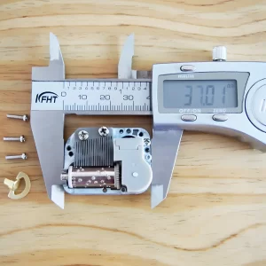 A digital caliper measuring the length of the mini music box mechanism, showing a reading of 37.01 mm. Three screws and a winding key are also visible.