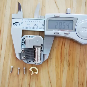 A digital caliper measuring the width of the mini music box mechanism, showing a reading of 28.54 mm. Three screws and a winding key are also visible.