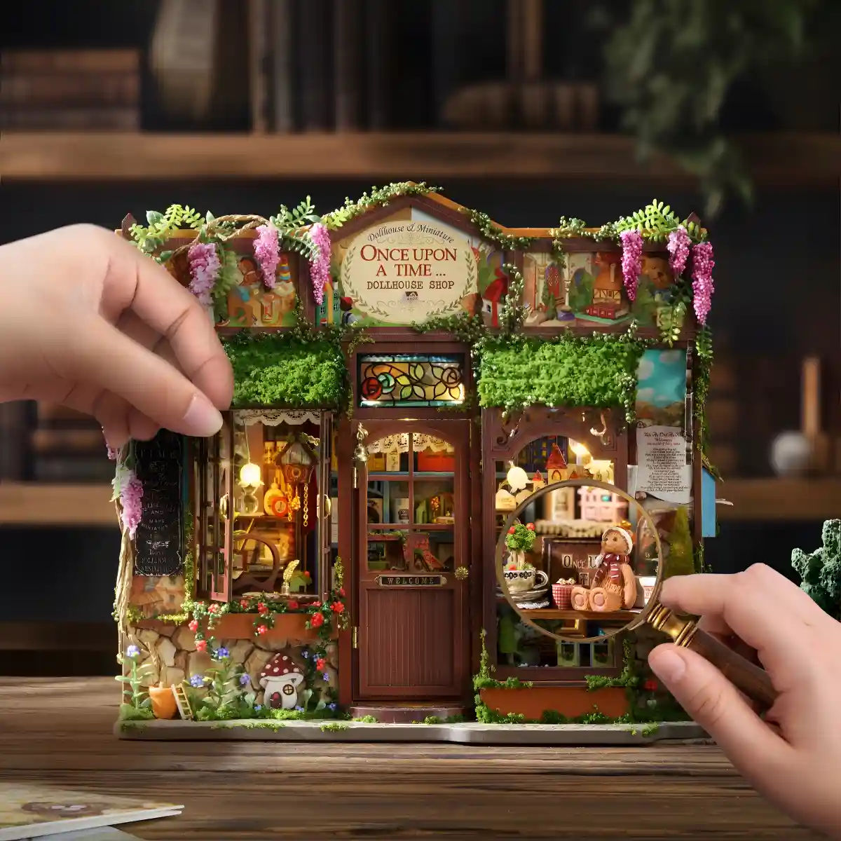 A person's hands holding a magnifying glass to a window of the miniature dollhouse, focusing on a tiny teddy bear and other objects inside the shop.