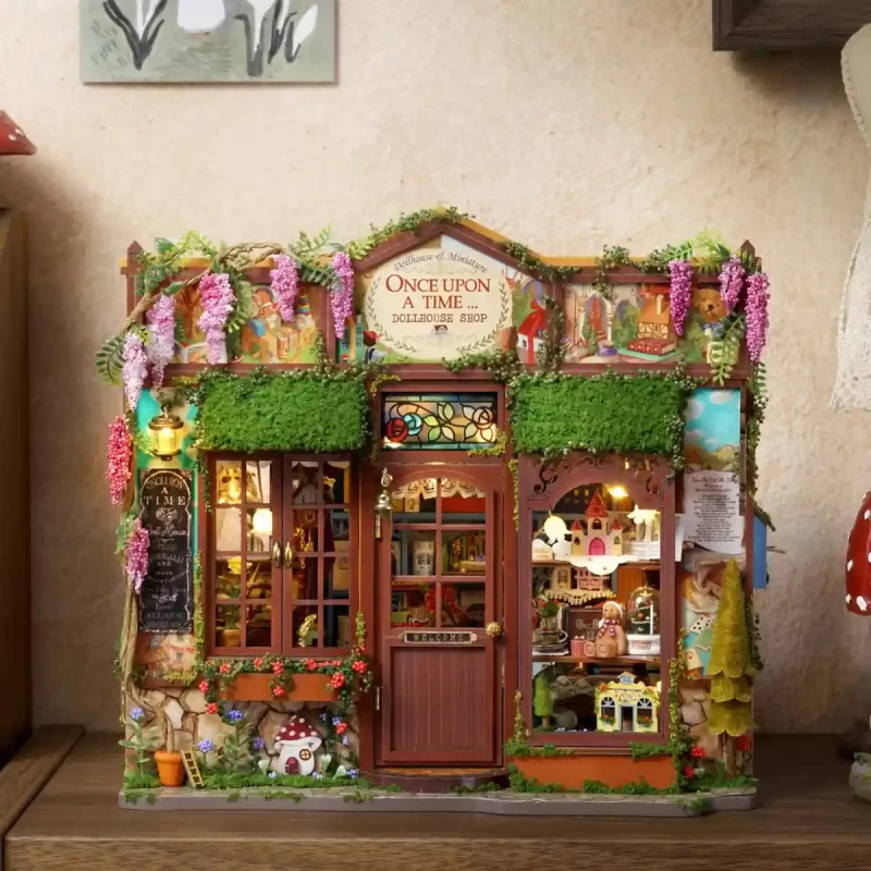 A straight-on view of the miniature dollhouse storefront, featuring a detailed door, a stained-glass window, overgrown vines with pink flowers, and a blackboard sign on the left.