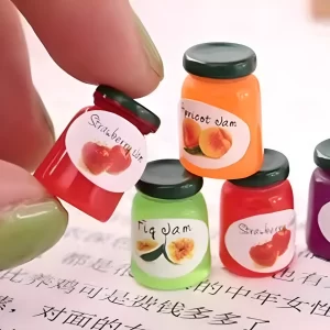 A hand holding a tiny strawberry jam jar miniature with other dollhouse jam jars.