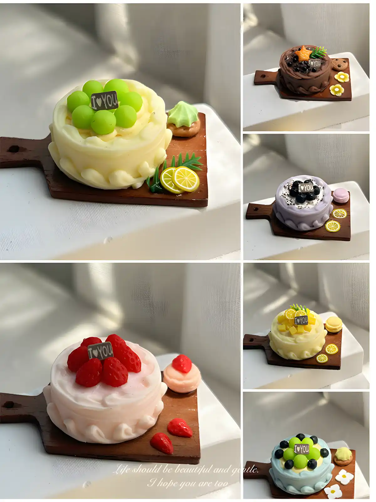 Six miniature cakes are arranged in two columns. The cakes have various toppings like green grapes, strawberries, chocolate, and yellow fruit slices. Each is on a small wooden board.