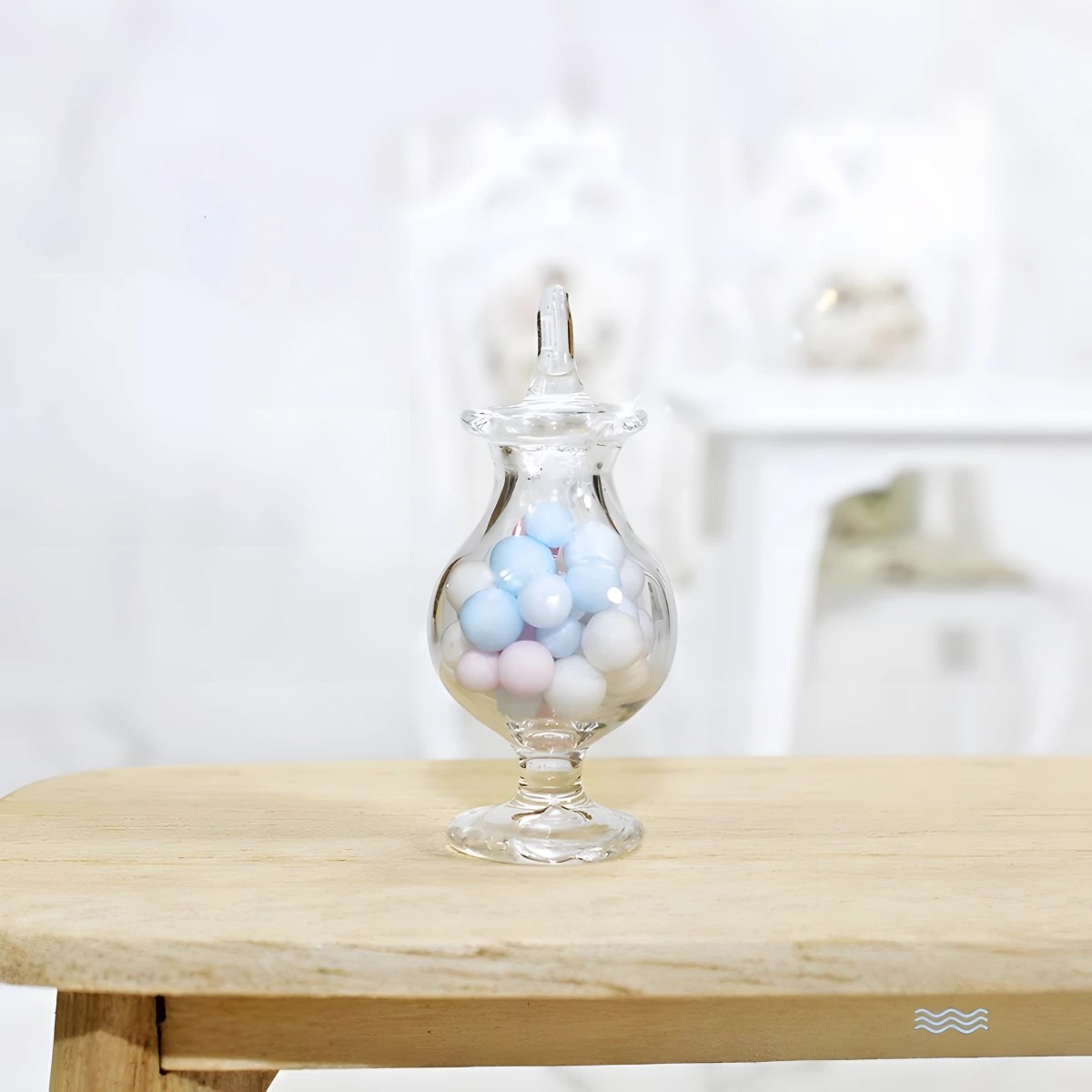 A miniature glass candy display jar, filled with light blue, pink, and white candies, stands on a wooden table in a dollhouse setting.