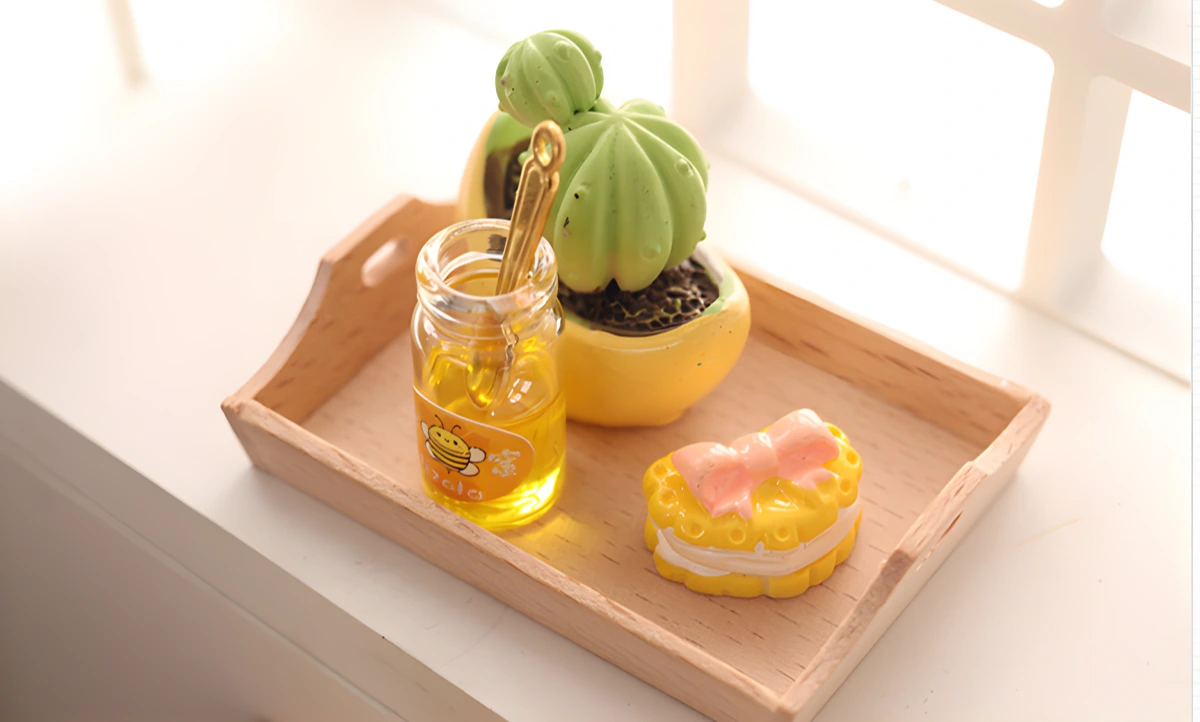 A miniature honey jar with a bee label and a honey dipper, a tiny potted cactus, and a small yellow cake on a wooden tray on a windowsill.
