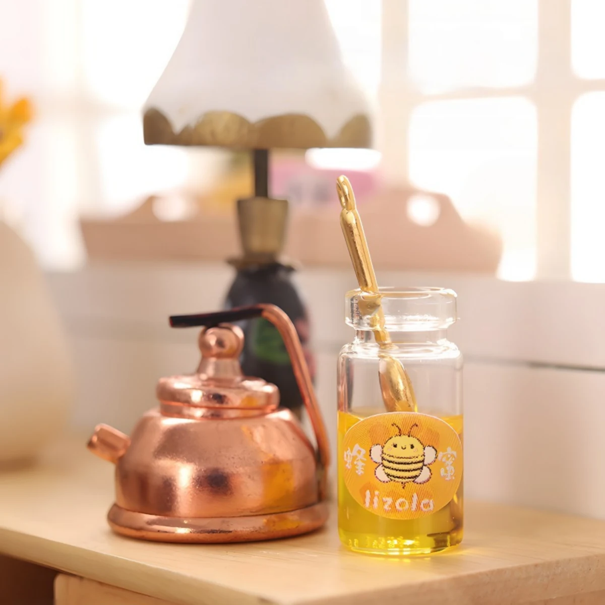 A miniature honey jar with a bee label and a honey dipper inside, sitting next to a tiny copper kettle on a wooden dollhouse table.