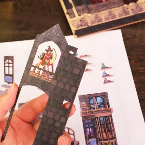 A hand holding a vibrant, pre-cut wooden piece of a witch for the Magic Alley DIY Book Nook Kit, showcasing the easy-to-assemble parts.